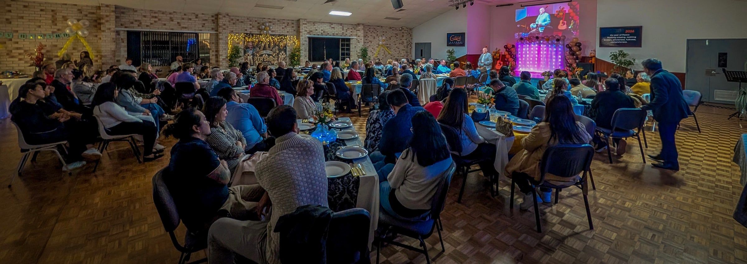 A full hall of people from many backgrounds seated at tables during a church gathering at CoastWIDE Church, Arundel, the stage lit pink