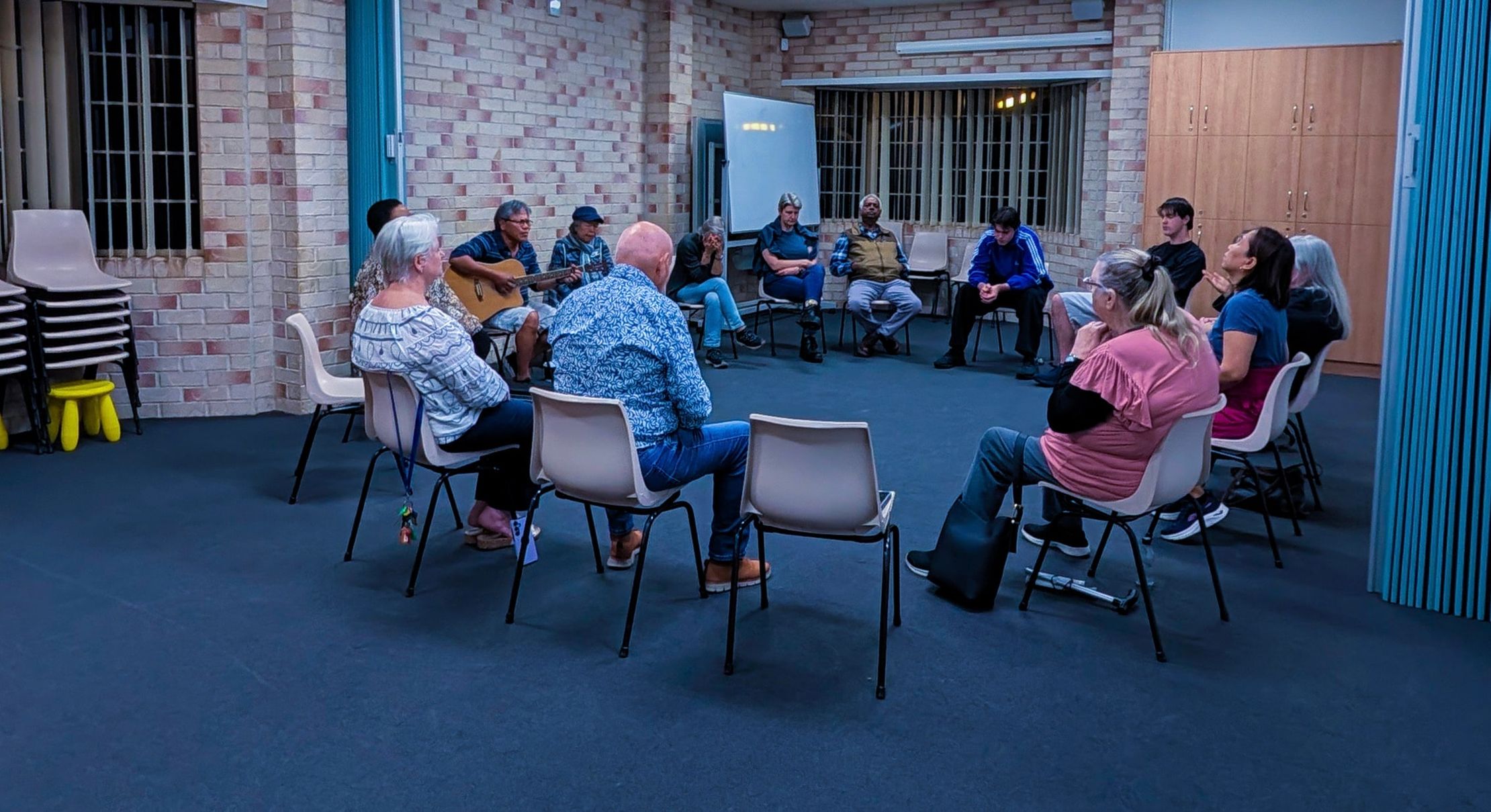 A circle of people seated in chairs pray together at the Wednesday prayer meeting in the hall at CoastWIDE Church, Arundel