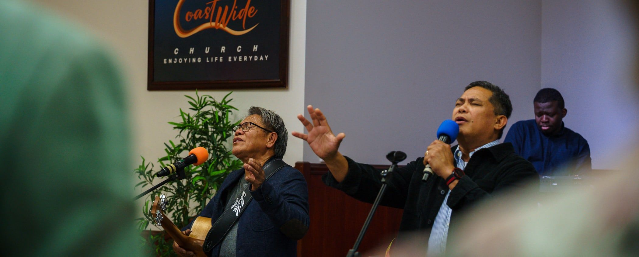 Pastor Herane Suralta leads Sunday worship beside the lit CoastWIDE Church sign, with Joel Gonzales on guitar