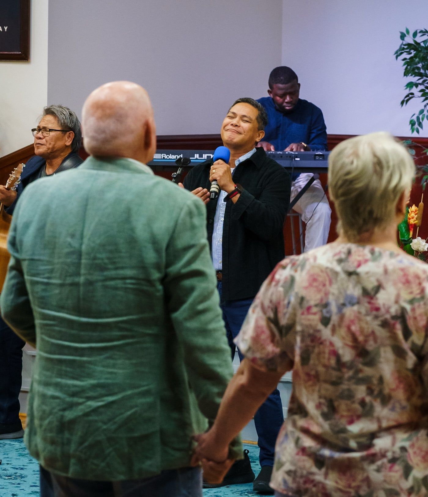 The congregation holding hands in worship while Pastor Herane leads with a microphone