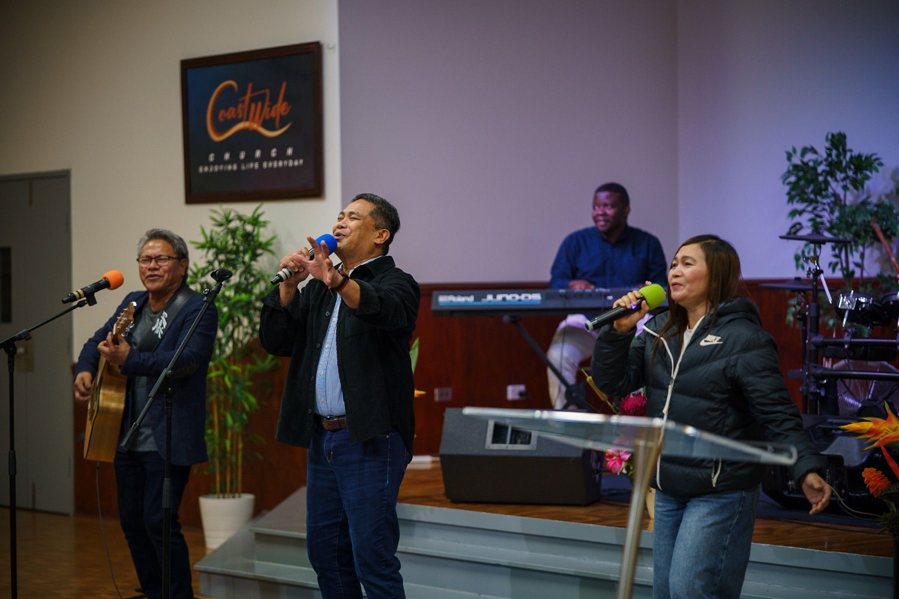 Pastor Herane Suralta leads worship with Joel Gonzales on guitar beside the CoastWIDE Church logo sign in Arundel