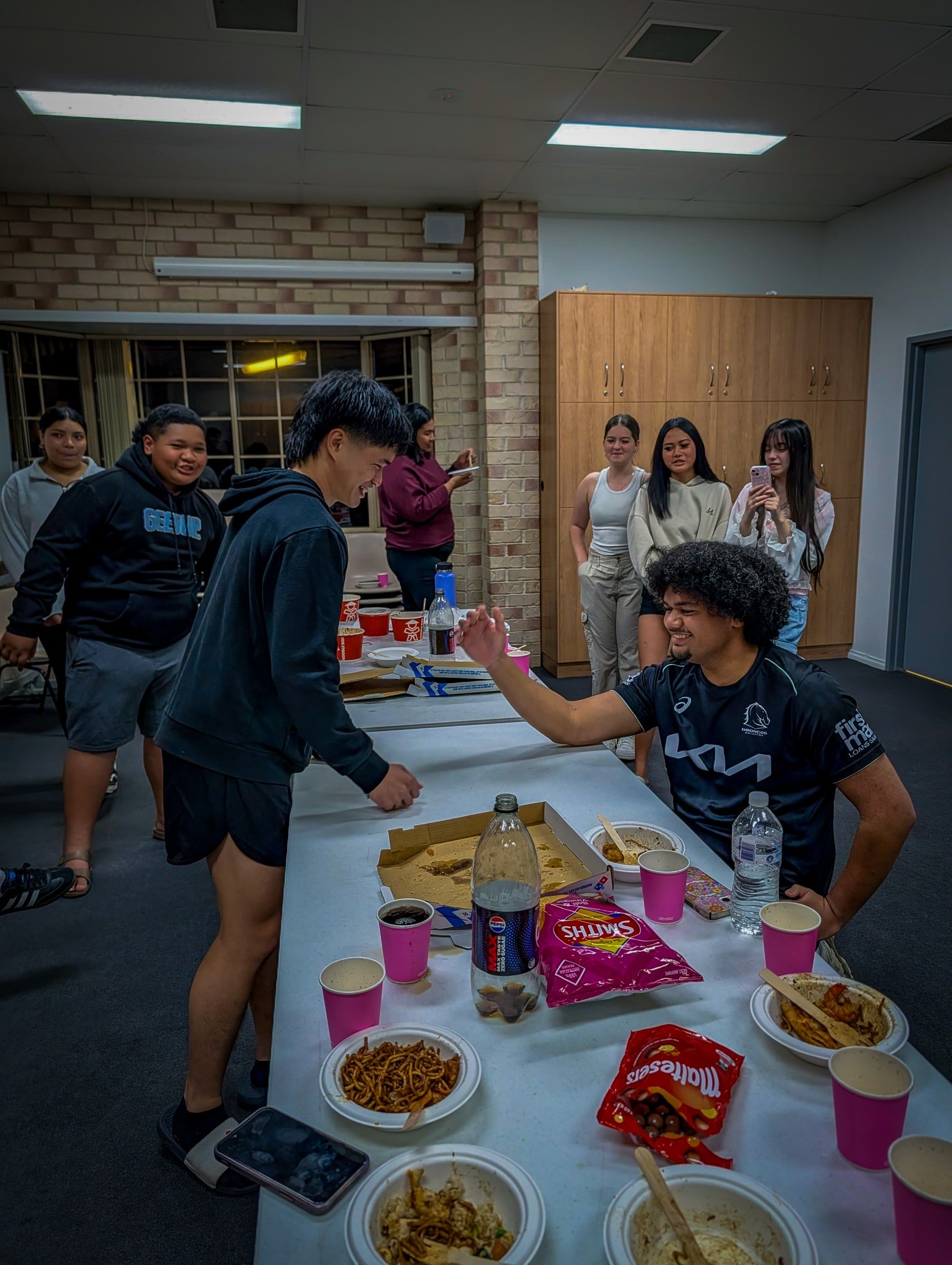 Young adults laughing and sharing food around a long table at a CoastWIDE Church Young Adults night in Arundel