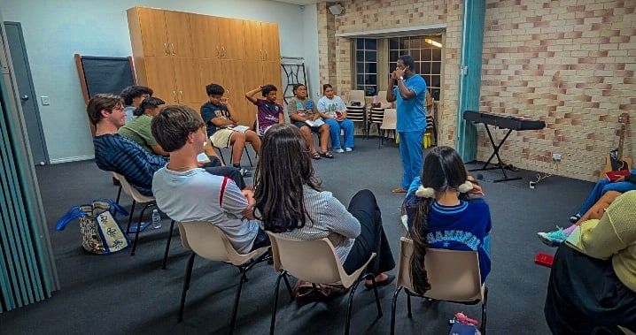 Young adults seated in a circle of chairs listen to a speaker standing in the middle of the room at a CoastWIDE Church Young Adults night in Arundel
