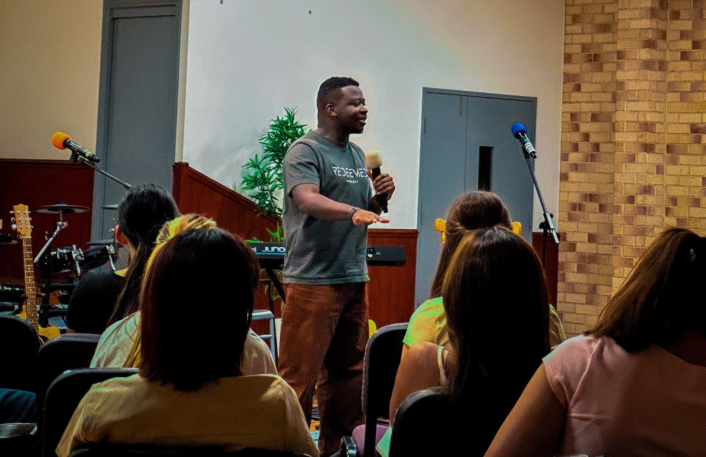 Young Adults Leader Delight Madujibeya speaks with a microphone to young adults seated in rows at CoastWIDE Church, Arundel
