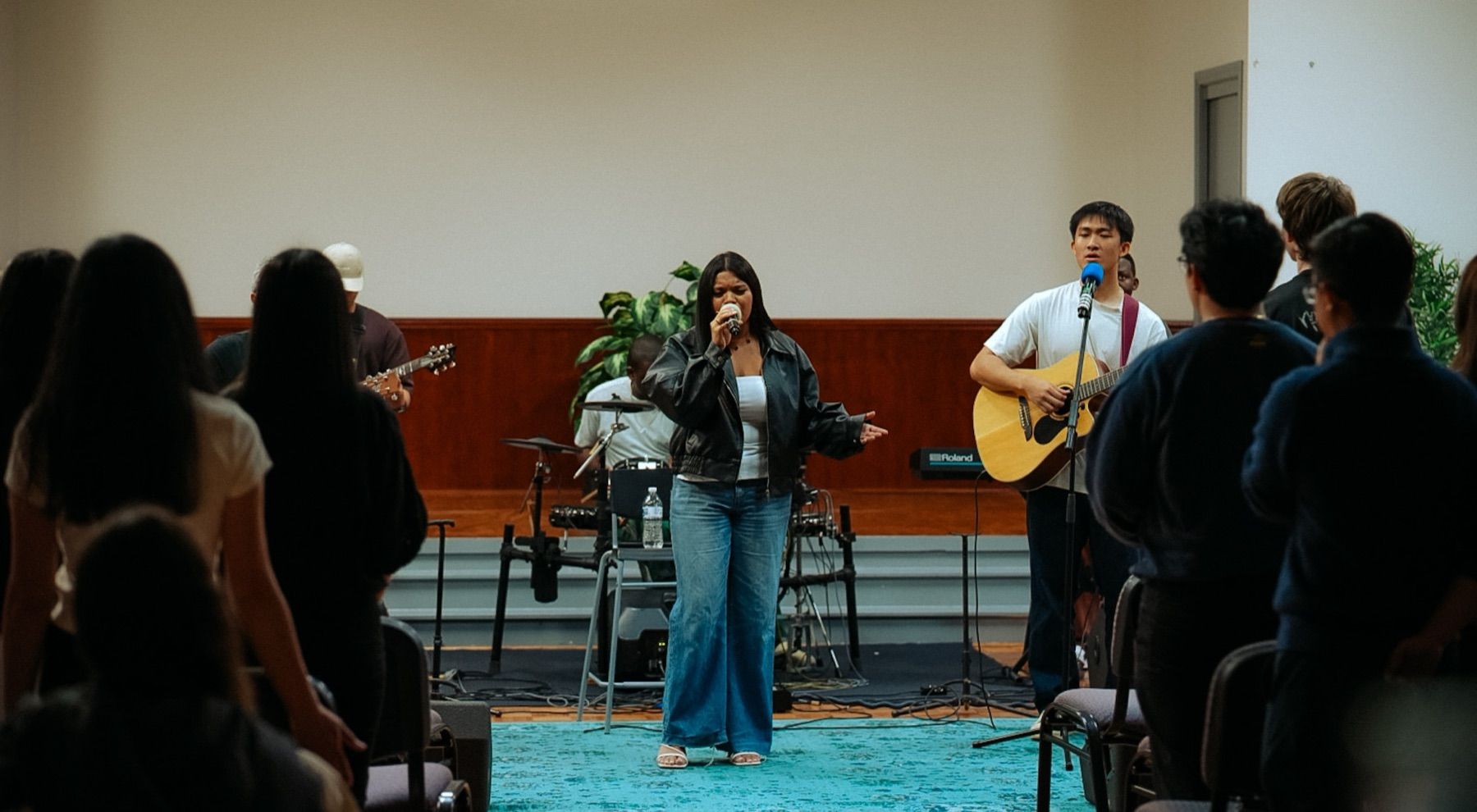 A young woman sings on stage beside a guitarist as young people stand to worship