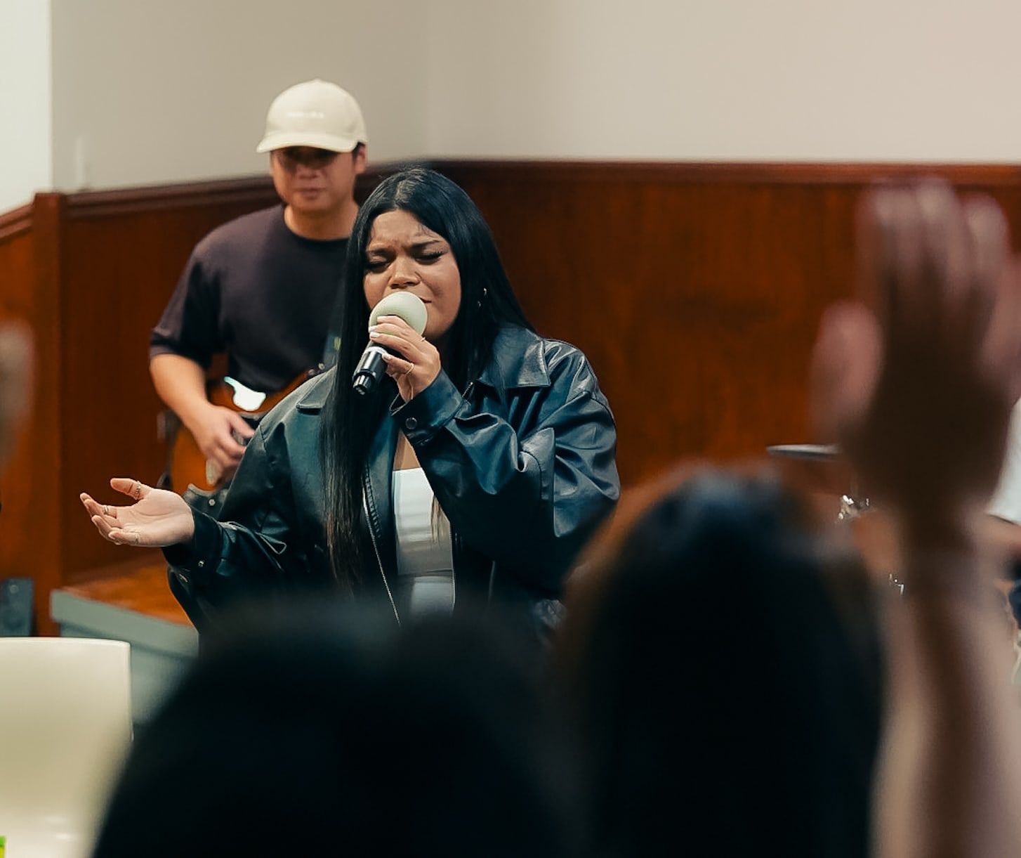 A young woman sings into a microphone with her hand outstretched during worship at CoastWIDE Church, a guitarist behind her and hands raised in the room